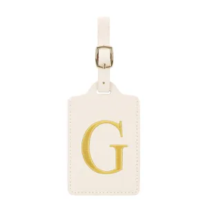 Vue détaillée d'une étiquette de bagage en similicuir beige avec lettre G brodée dorée.