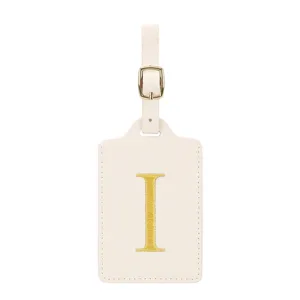 Gros plan détail d'étiquette bagage beige avec lettre brodée dorée I et boucle en métal.