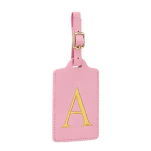 Dettaglio etichetta bagaglio in pelle PU rosa con lettera ricamata dorata e fibbia in metallo.