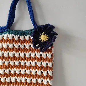 Handgemaakte gehaakte shopper met blauwe handvatten en bloem, stijlvol gestreept ontwerppatroon.