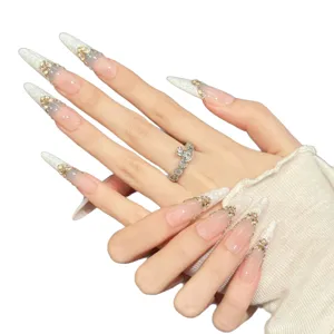 Ongles stiletto extra longs avec dégradé nude, texture blanche et strass papillon dorés.