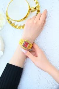 Détail de montre à quartz carrée dorée avec cadran rouge vif et chiffres romains.