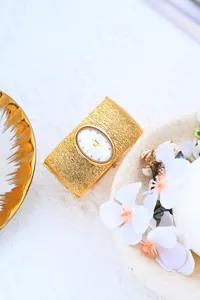 Detalle de reloj pulsera rígida dorada, correa texturizada, elegante esfera blanca ovalada.