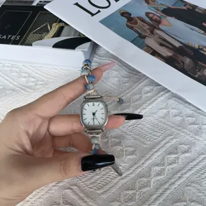 Nahaufnahme einer minimalistischen Quarzuhr mit eckigem Zifferblatt und Perlenarmband.