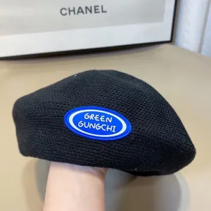 Close-up detail of a stylish black knit beret featuring a blue oval embroidered logo patch.