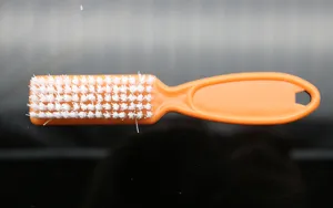 Orange plastic nail cleaning brush with white hard bristles and tail hanging hole detail view.