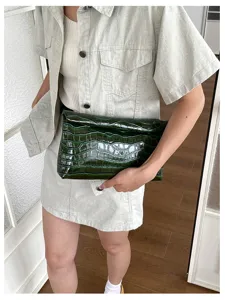 Close-up view of the dark green glossy crocodile embossed leather clutch bag held by model.