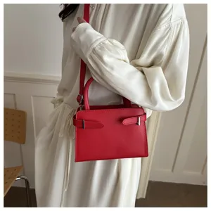 Detailed shot of a stylish red crossbody bag with metal buckle straps worn by a model in white dress.
