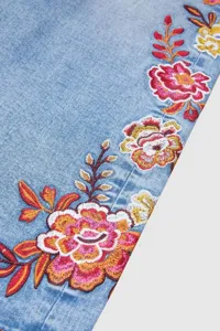 Vista detallada de vaqueros de mujer en denim azul con bordado floral colorido.