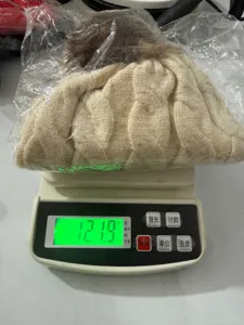 Detailed weight measurement of a beige chunky knit beanie with fur pom-pom on a scale