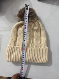 Cream knit beanie with brown fur pom-pom shown with a measuring tape for height