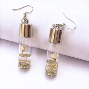 Close up of clear glass bottle earrings filled with tiny white flowers, liquid and gold glitter.