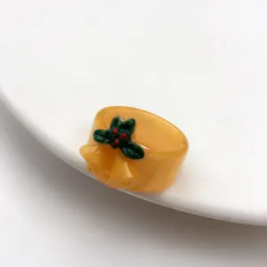Anello in resina natalizia con agrifoglio verde e campanella, dettaglio di gioielli festivi.