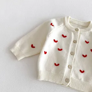 Vista dettaglio cardigan in maglia per neonati, ricamo cuore rosso e bottoni su tessuto bianco.