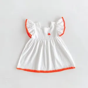 White baby girl dress with orange pom-pom trim and flutter sleeves. Cute summer toddler outfit detail.
