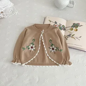 Detail shot of baby knit cardigan with floral embroidery and ruffle trim on elegant lace tablecloth.