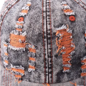 Detail view of distressed denim baseball cap with contrast orange stitching and ripped holes texture.