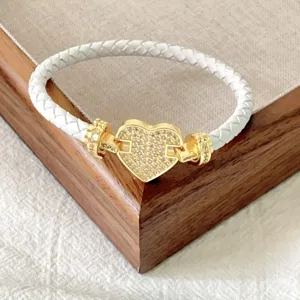 Dettaglio bracciale in pelle intrecciata bianca con chiusura a cuore dorata su vassoio in legno.