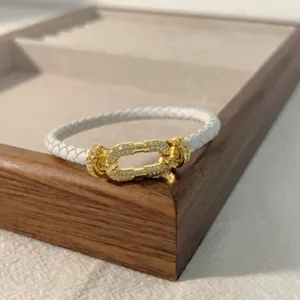 White braided leather bracelet with gold horseshoe clasp, detailed view of micro-pave crystal link.