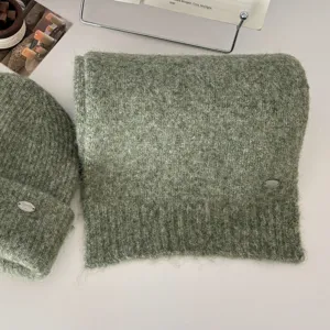 Vista detallada del conjunto de bufanda y gorro de punto verde suave con elegante placa metálica.