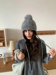 Detalle del gorro de punto gris con pompón acogedor y cordones trenzados para orejeras.