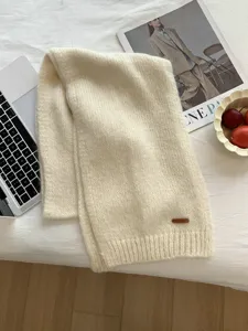 Detailaufnahme eines weichen, beige Strickschals mit Lederetikett auf weißem Bett.