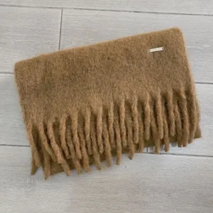 Detailaufnahme des braunen Winterschals mit dicken Fransen und kleinem Metalllogo.