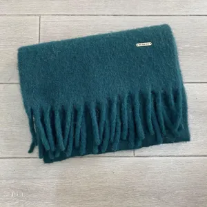 Detailansicht des dunkelgrünen Winter Fransenschals mit feinem Metalllogo aus weichem Stoff.