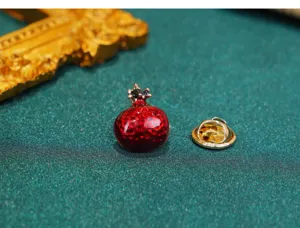 Close-up of a miniature red pomegranate brooch with glossy enamel and gold-toned backing.