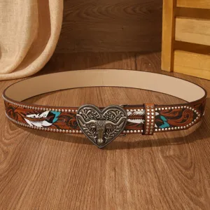 Detail view of the western leather belt featuring a carved bull skull heart buckle and feather motifs.