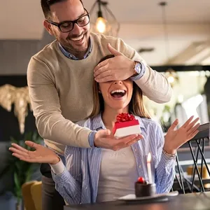 Lifestyle scene showing a man surprising a woman with a gift box during a birthday celebration.