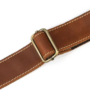 Leather bag strap detail with bronze metal buckle and white stitching, brown cowhide.