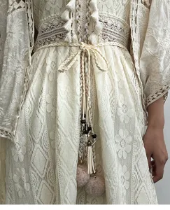 Gros plan d'une robe en dentelle crème bohème détaillée avec une ceinture tressée et des pompons.