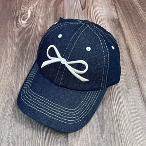 Gros plan détaillé de la casquette de baseball en jean bleu foncé brodée d'un nœud blanc.
