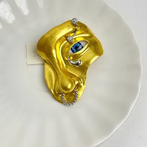 Surreële gouden gezichtsbroche met realistisch blauw oog en unieke piercingdetails.