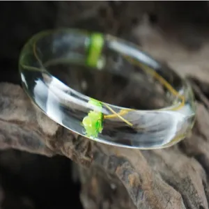 Clear resin bracelet detail showing embedded green dried flowers on a rustic wooden background.