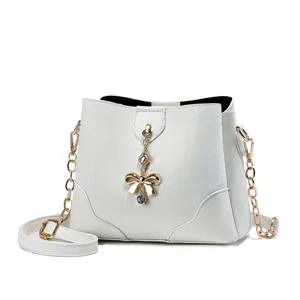 Elegante borsa a secchiello bianca con catena dorata, ciondolo con fiocco e design chic.