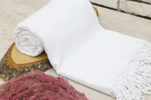 White Turkish beach towel with fringe, rolled up on wood display with pink floral decor.