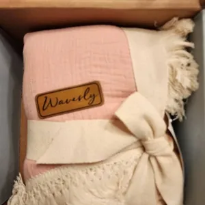 Close-up of pink muslin baby swaddle blanket detail featuring soft fringe and branded leather tag.
