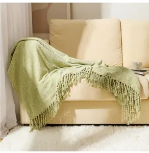 Green textured chenille throw blanket with elegant tassels draped on a cozy beige couch.