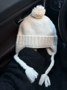 Cozy white knitted earflap beanie hat with cute pom pom and braided tassels for winter warmth.