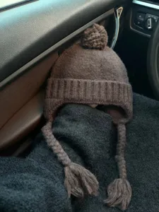 Brown knitted earflap hat with pom-pom and braided straps, warm and windproof winter beanie.