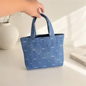 Primer plano del minibolso tote de mezclilla azul con bordado de lazo blanco, sostenido a mano.