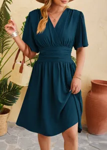 Detail view of dark teal V-neck dress with pleated waist and flutter sleeves in a scenic setup.