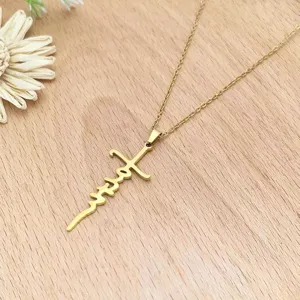 Close-up detail of a gold stainless steel cross necklace with cursive script pendant on wood.