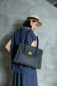 Detail shot of black leather shoulder bag with gold hardware worn by model against grey wall.