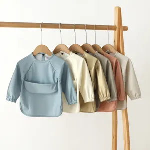 Waterproof long sleeve baby smock bib with food catcher pocket, hanging on wooden rack detail view.