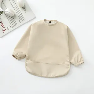 Vista detallada del babero impermeable beige para bebés, con mangas largas y bolsillo recogemigas.
