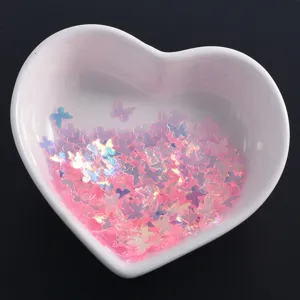 Close-up of holographic pink butterfly nail sequins displayed in a white heart bowl.