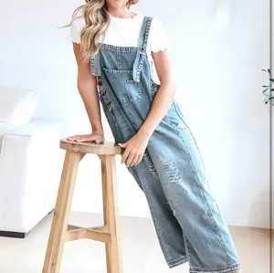 Detailed view of distressed denim overalls paired with white tee, posing near wooden stool.